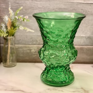 Könnte beinhalten: Eine grüne Glasvase mit strukturiertem Muster. Die Vase hat eine weite Öffnung und einen bauchigen Boden. Sie steht auf einer weißen Oberfläche. Im Hintergrund befindet sich eine kleinere Glasvase mit Blumen. Die Vase ist etwa 20 cm hoch.