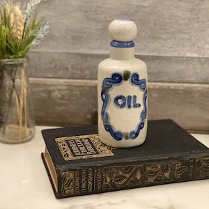 Puede incluir: Una botella de aceite de cerámica blanca con detalles decorativos azules y verdes y la palabra "OIL" en el centro. La botella tiene una parte superior redondeada y está sobre un libro antiguo.