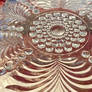 May include: A clear glass plate with a decorative design of a circle of raised dots surrounded by a leafy pattern.