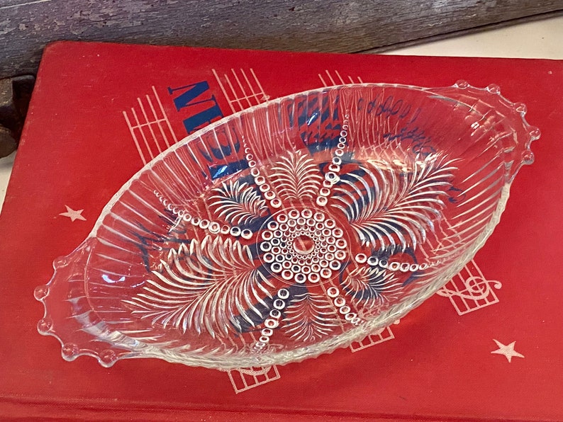 May include: A clear glass serving dish with a scalloped edge and a decorative pattern of leaves and circles. The dish is designed for serving snacks or small desserts.