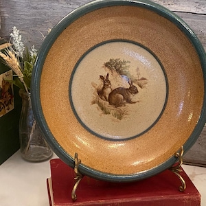 May include: A decorative ceramic bowl with a tan and teal rim. The center features a detailed illustration of two rabbits in a natural setting. The bowl is displayed on a gold-colored stand.