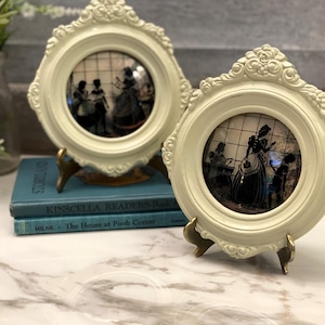 Puede incluir: Dos marcos decorativos redondos de color crema con bordes ornamentados y grabados florales. Cada marco contiene una ilustración circular de siluetas que representa figuras en una habitación, sobre soportes de metal dorado.