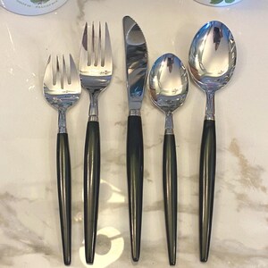 Puede incluir: Un juego de cinco utensilios de acero inoxidable: un tenedor, un cuchillo, una cuchara y dos tenedores más pequeños. Cada pieza tiene una cabeza plateada brillante y un mango negro. Los utensilios están dispuestos sobre una superficie clara, similar al mármol.