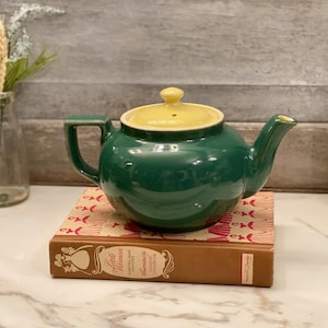May include: A green ceramic teapot with a yellow lid sits atop a book. The teapot has a rounded body, a curved spout, and a handle. The book is brown with red and white accents. A glass vase with flowers is in the background.