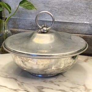 Puede incluir: Un cuenco de metal plateado con tapa. El cuenco tiene forma redonda con un borde decorativo con cuentas y un asa en la tapa. El cuenco está sobre una superficie de mármol blanco.