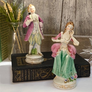 May include: Two porcelain figurines depicting a person in a purple coat with yellow striped breeches and a person in a pink bodice with a green skirt. Both figures stand on circular bases with gold trim, featuring painted details and glossy finishes.