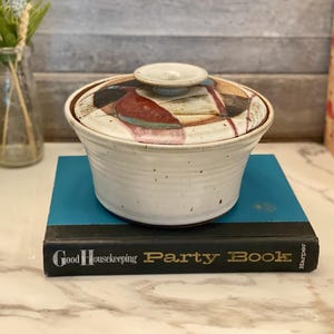 Puede incluir: Una fuente de cerámica con tapa. El plato es blanco con ribete marrón y una tapa con un diseño abstracto colorido. El plato está sobre un libro azul con el texto "Good Housekeeping Party Book".