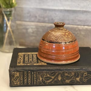 May include: A handcrafted ceramic jar with a lid. The jar features a warm color palette of brown and orange, with a decorative wavy pattern on the lid. The jar is sitting on a vintage book.