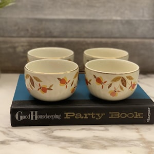 Può includere: Quattro ciotole color crema con bordi dorati, decorate con un motivo floreale marrone, arancione e rosa. Le ciotole sono disposte su un libro blu intitolato "Good Housekeeping Party Book".