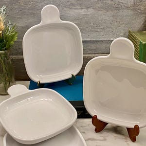 May include: A collection of white ceramic serving dishes. Each dish has a square shape with rounded corners and a handle. The dishes are displayed on small wooden and metal stands, showcasing their design.