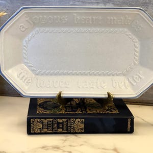May include: A white rectangular serving platter with a blue trim and the words "a joyous heart makes the loaves taste better" embossed on the surface. The platter is displayed on a dark blue book with gold lettering.