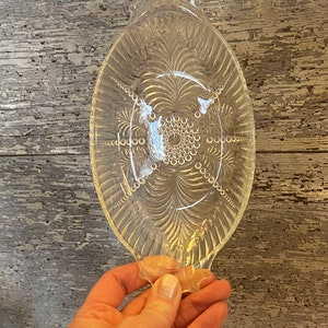May include: A clear glass serving dish with a scalloped edge and a decorative pattern of circles and lines. The dish is oval shaped and has a handle on each end.