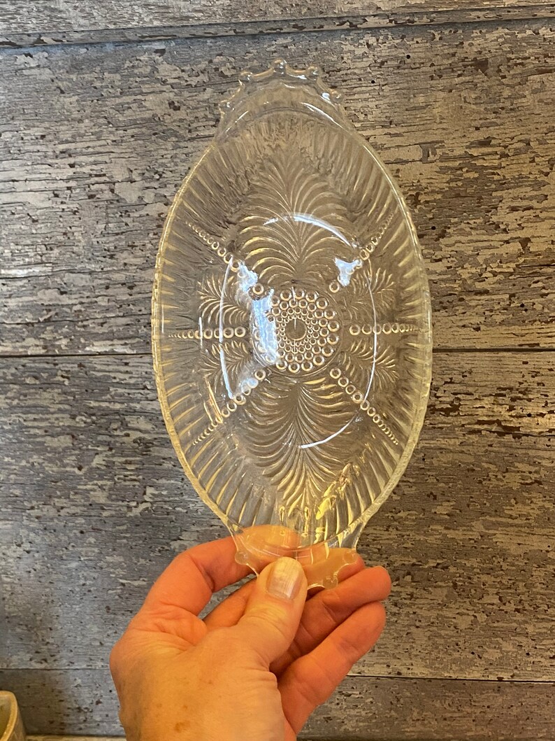 May include: A clear glass serving dish with a scalloped edge and a decorative pattern of circles and lines. The dish is oval shaped and has a handle on each end.