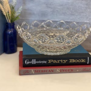 Puede incluir: Un cuenco de vidrio transparente con un patrón tejido decorativo. El cuenco está colocado encima de una pila de tres libros. El libro superior es negro con texto blanco que dice "Good Housekeeping Party Book". El libro del medio es rojo con texto blanco que dice "The American Singer Book".