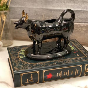 May include: Black ceramic creamer in the shape of a cow with gold accents. The creamer is sitting on a book with a green cover and gold lettering.