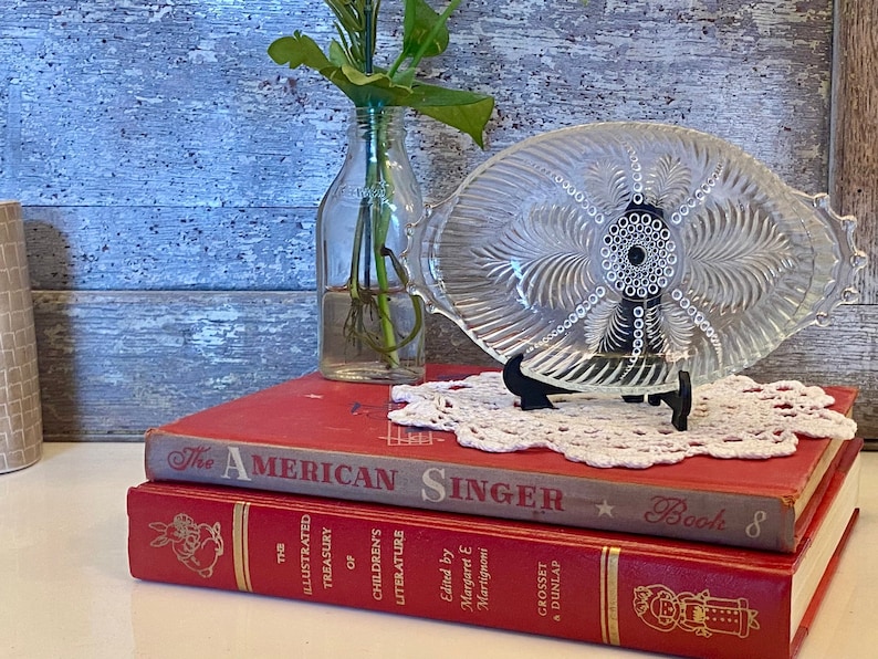 May include: A clear glass serving dish with a decorative etched pattern sits on a white doily atop two red books. The top book is titled "The American Singer, Book 8".