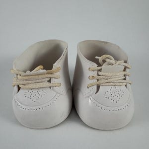 Op de afbeelding: Een paar witte poppenschoenen met een gladde afwerking, ronde neuzen en een geperforeerd hartvormig patroon op de wreef. De schoenen zijn voorzien van beige stoffen veters door oogjes, met een lage enkelopening en een stevige constructie.
