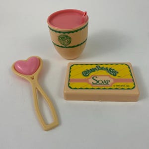 May include: A collection of vintage Cabbage Patch Kids toys, including a pink and tan cup, a heart-shaped rattle, and a bar of soap with the words "Soap" and "Cabbage Patch Kids" printed on it.
