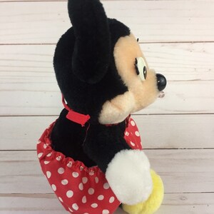 May include: A plush black and white Minnie Mouse toy wearing a red dress with white polka dots. The toy is sitting on a white wooden surface.