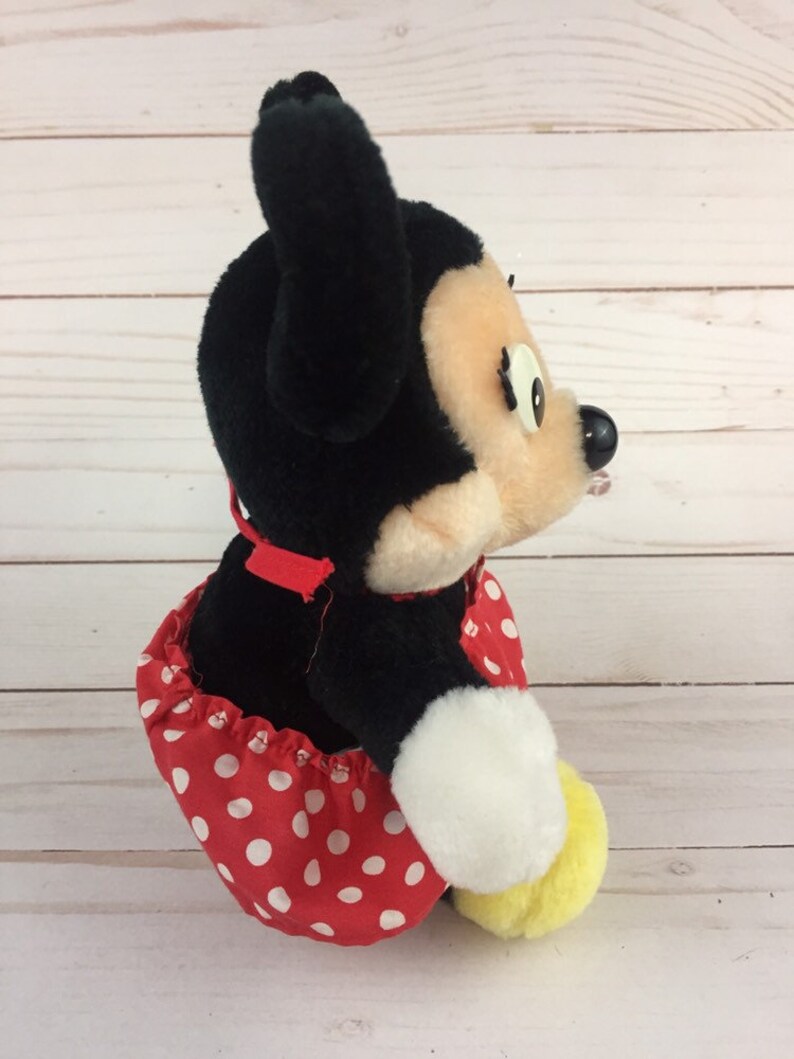May include: A plush black and white Minnie Mouse toy wearing a red dress with white polka dots. The toy is sitting on a white wooden surface.