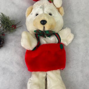 May include: White plush Christmas stocking shaped like a bear wearing a Santa hat and a green and red plaid bow tie. The stocking is red and has a white trim.
