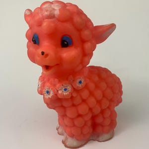 May include: A vintage, coral-colored lamb figurine with a textured body and blue-eyed expression. The lamb wears a floral collar with white and blue accents. Its hooves are a faded white color.