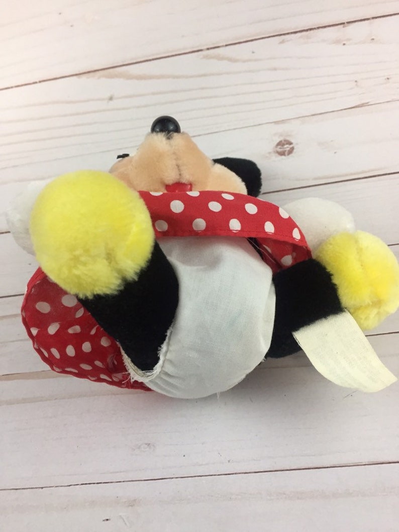 May include: A plush toy of Mickey Mouse wearing a red and white polka dot dress. The toy is lying on its back with its legs in the air.