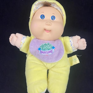 Peut inclure: Une poupée vintage Cabbage Patch Kids Babyland. La poupée porte une combinaison jaune clair avec une capuche assortie et un bavoir violet. La poupée a les yeux bleus et une expression souriante.