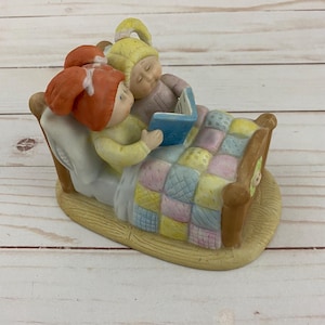 Op de afbeelding: Een keramieken beeldje met twee kinderen in bed die een boek lezen. De kinderen dragen nachtmutsen en zijn bedekt met een patchwork quilt. Het bedframe is bruin en de basis is beige.
