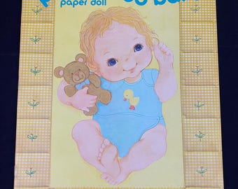 May include: A vintage paper doll book featuring a baby doll with blonde hair and blue eyes. The doll is holding a teddy bear and wearing a blue onesie with a yellow duck design. The book cover is yellow with a checkered pattern and the title "peek-a-boo baby paper doll" is printed in black.