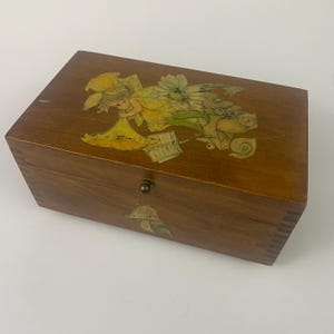 May include: Wooden box with a hinged lid and a brass clasp. The box is decorated with a colourful illustration of a fairy with yellow and green accents. The box has a natural wood grain finish.