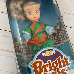May include: A vintage Bright Eyes doll wearing a green and orange striped dress with a red bow. The doll has blonde hair and blue eyes. The doll is packaged in a box with the words "Taffy Twis" and "Bright Candy Stripe Doll" on it.