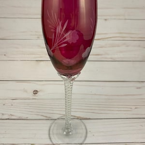 May include: A tall, clear glass champagne flute with a twisted stem. The bowl of the flute is a deep, rich pink color with a white etched floral design.