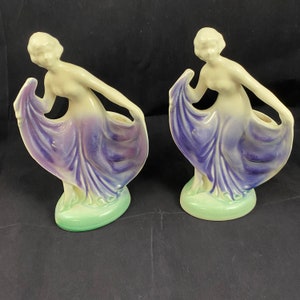 Puede incluir: Par de figuritas de cerámica vintage de mujeres con vestidos morados fluidos. Las figuritas están en posición de pie y tienen una estética delicada y femenina.
