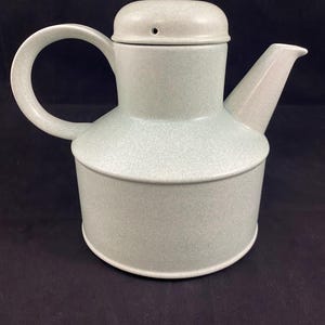 Op de afbeelding: Een lichtgrijze theepot van keramiek met een rond lichaam en een deksel met een klein gaatje. De theepot heeft een handvat en een tuit.