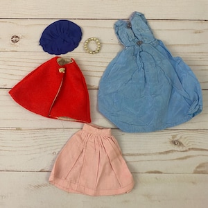 May include: A collection of doll clothes, including a blue dress, a red cape, a pink skirt, a blue hat, and a gold beaded necklace. The doll clothes are arranged on a light wood surface.