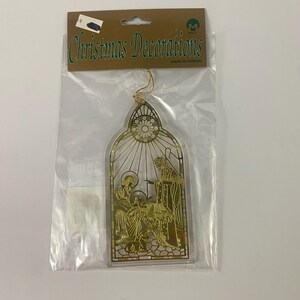 May include: Gold-colored Christmas decoration in a clear plastic bag. The ornament features a religious scene with figures and the words "Christmas Decorations". The ornament is shaped like a stained-glass window.
