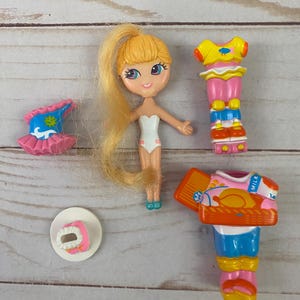 May include: A small doll with blonde hair in a ponytail, wearing a white swimsuit, and several outfits. The outfits include a pink and blue tutu, a yellow top with pink pants, and a pink and orange outfit with a basket.