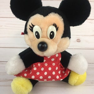 May include: A plush Minnie Mouse toy wearing a red dress with white polka dots. She has black ears, a black nose, and large white eyes with eyelashes.