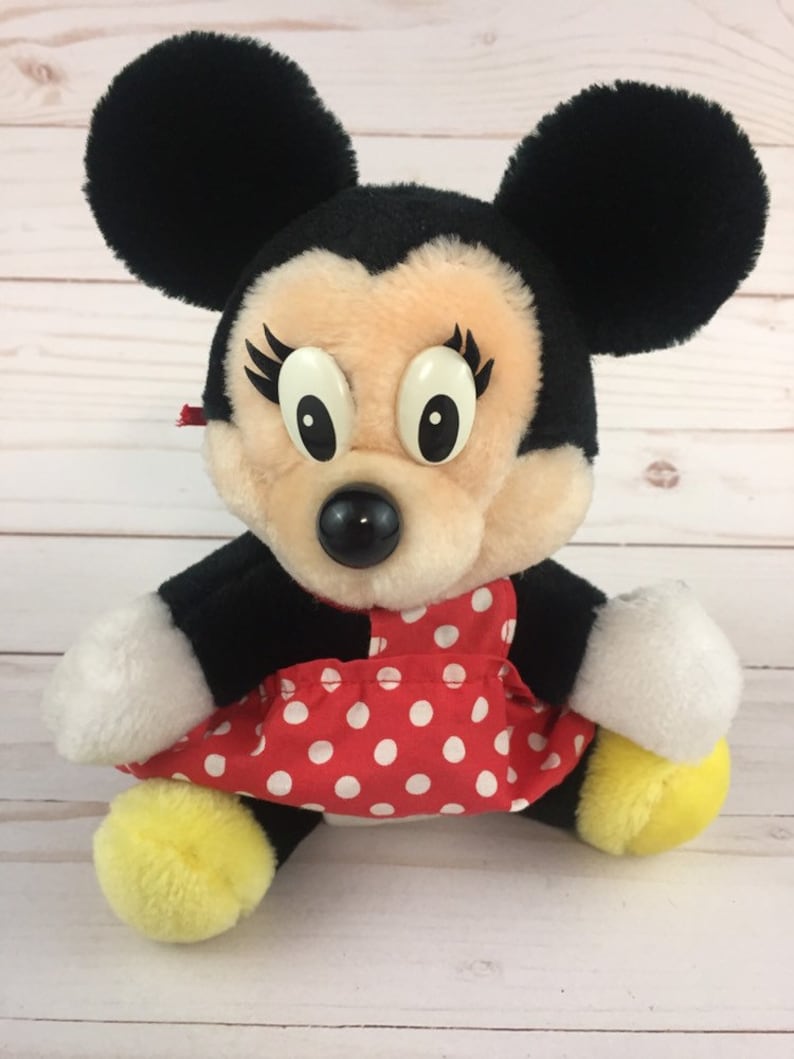 May include: A plush Minnie Mouse toy wearing a red dress with white polka dots. She has black ears, a black nose, and large white eyes with eyelashes.