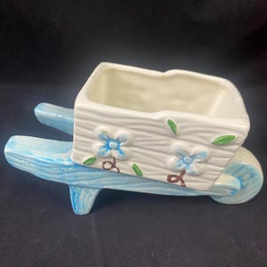 May include: A blue and white ceramic wheelbarrow planter with a floral design. The planter has a rectangular shape and is decorated with blue flowers and green leaves.