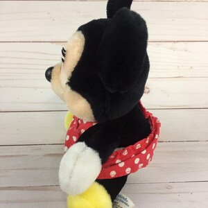 May include: A red and white polka dot dress with a yellow bow on the back. The dress is on a black and white plush Mickey Mouse toy. The toy has a black nose and big ears. The toy is standing on a white wooden surface. The tag on the toy says "Disney World".
