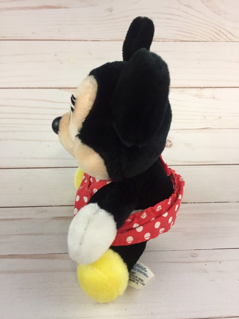 May include: A red and white polka dot dress with a yellow bow on the back. The dress is on a black and white plush Mickey Mouse toy. The toy has a black nose and big ears. The toy is standing on a white wooden surface. The tag on the toy says "Disney World".