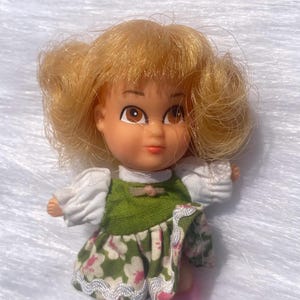 Könnte beinhalten: Eine Vintage-Puppe mit blonden Haaren und braunen Augen. Die Puppe trägt ein grünes Kleid mit Blumenmuster und weißen Ärmeln. Die Schuhe der Puppe sind rosa.
