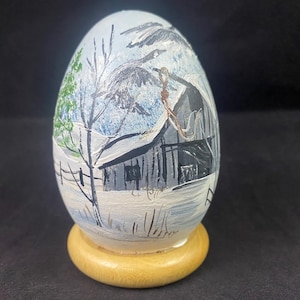 May include: A hand-painted egg with a winter scene. The egg depicts a white farmhouse with a snow-covered roof, trees, and a fence. The egg is sitting on a wooden base.