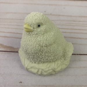 May include: A small, white ceramic bird figurine with a yellow beak and black eyes. The bird is sitting in a nest made of the same material.