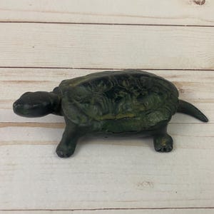 May include: A green cast iron turtle figurine. The turtle is in a walking position with its head turned to the left. The shell has a detailed design.