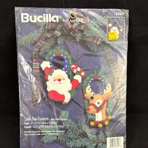 Pode incluir: Um kit de apliques de feltro Bucilla para maçanetas com o Pai Natal e o Rudolph. O pacote inclui feltro estampado, fio de bordar, lantejoulas, missangas e agulhas. As medidas finais são 17,78 cm x 22,86 cm e 12,07 cm x 24,13 cm.