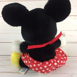 May include: A black and white plush Mickey Mouse doll wearing a red dress with white polka dots and a red bow. The doll is sitting on a white wooden surface.