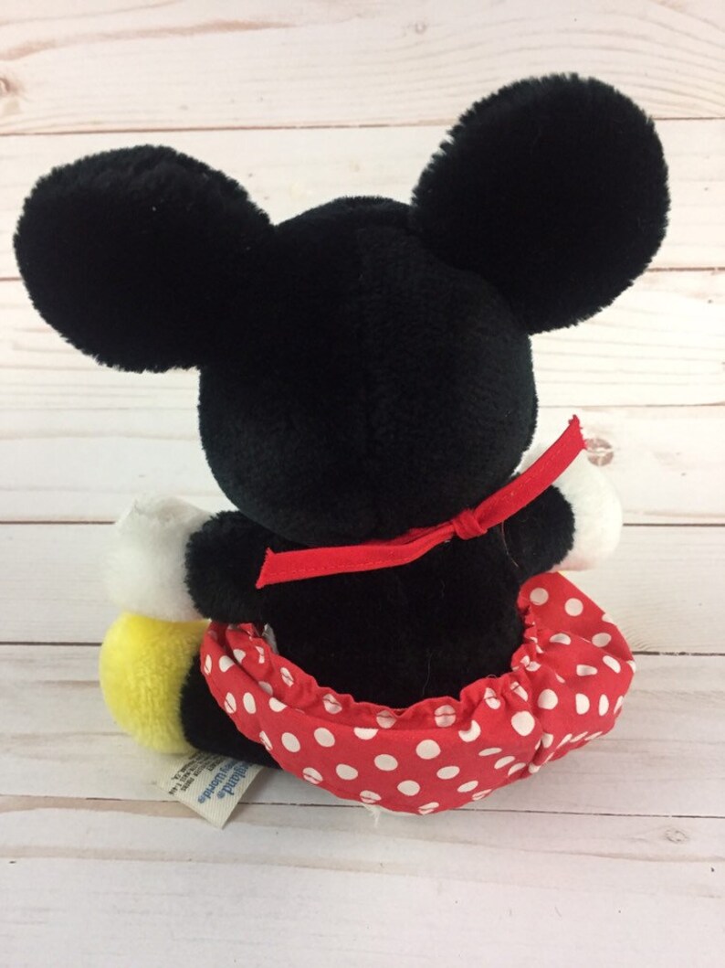 May include: A black and white plush Mickey Mouse doll wearing a red dress with white polka dots and a red bow. The doll is sitting on a white wooden surface.